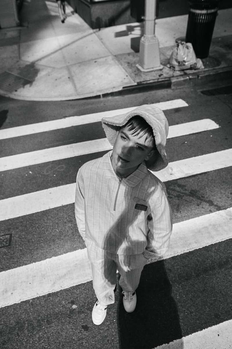 High angle view of a young adult in a sunhat and pinstripe jumpsuit standing on a crosswalk in a black and white street photography style.
