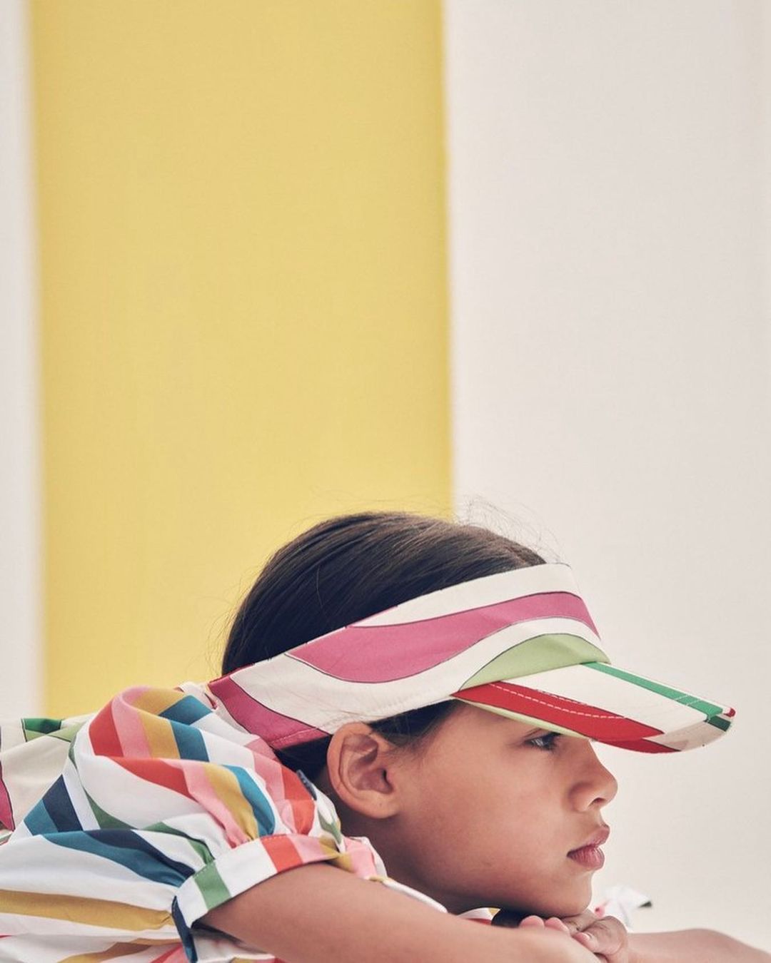 Child in colorful striped visor and shirt poses indoors against a yellow and white backdrop, resting chin on clasped hands.