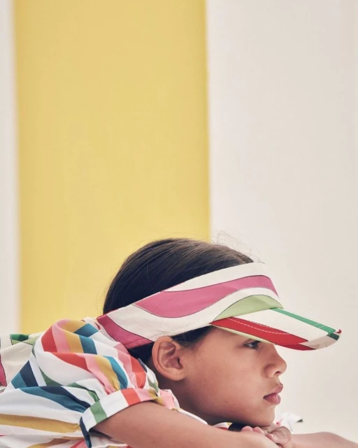 Child in colorful striped visor and shirt poses indoors against a yellow and white backdrop, resting chin on clasped hands.