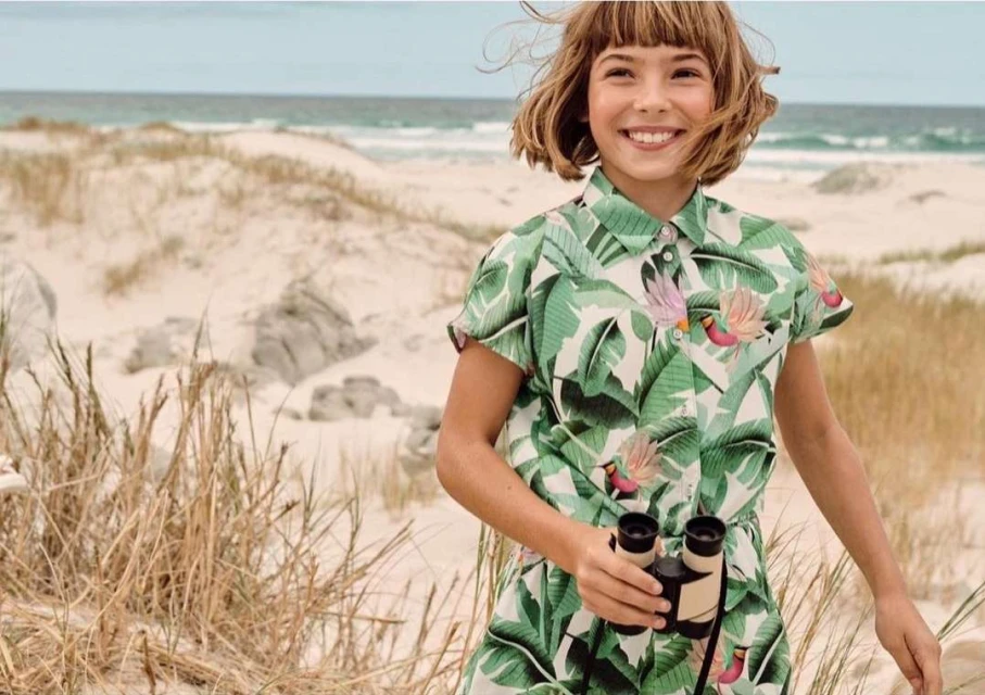 A girl smiles on a beach wearing a tropical leaf-patterned outfit, holding binoculars, with sand dunes and ocean visible in the background.