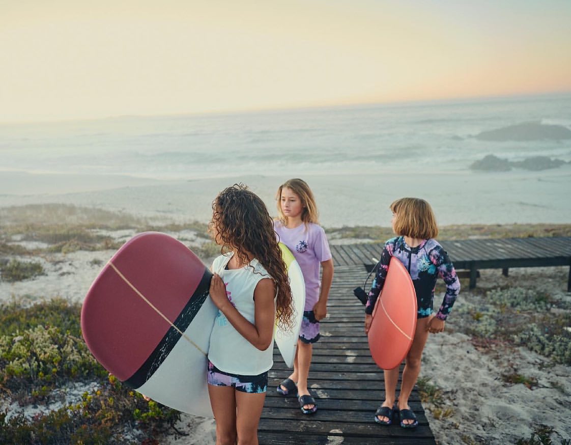 Three young girls with surfboards walk on a wooden path at the beach near the ocean on a sunny day at sunset.