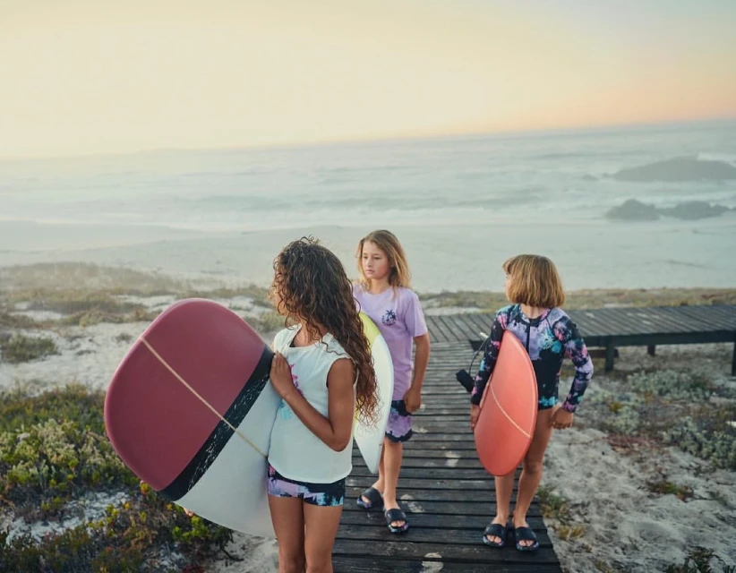 Three young girls with surfboards walk on a wooden path at the beach near the ocean on a sunny day at sunset.