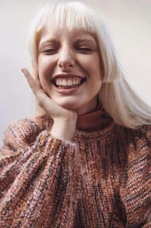 Close-up portrait of a laughing woman with blonde blunt bangs wearing a textured rust knit sweater, photographed by Franck Malthiery