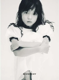 A young child wearing a white dress with dark ruffled trim poses against a plain white background in a black and white portrait.