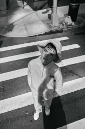 High-angle black and white street portrait of a young man wearing a bucket hat and striped coordinated set walking across a crosswalk