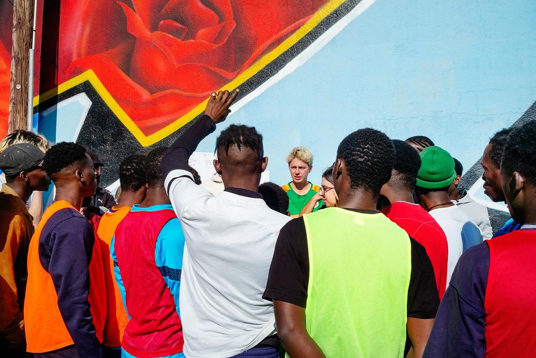 Group of people in colorful shirts gather around a person in a green vest in an outdoor urban setting with a mural background.