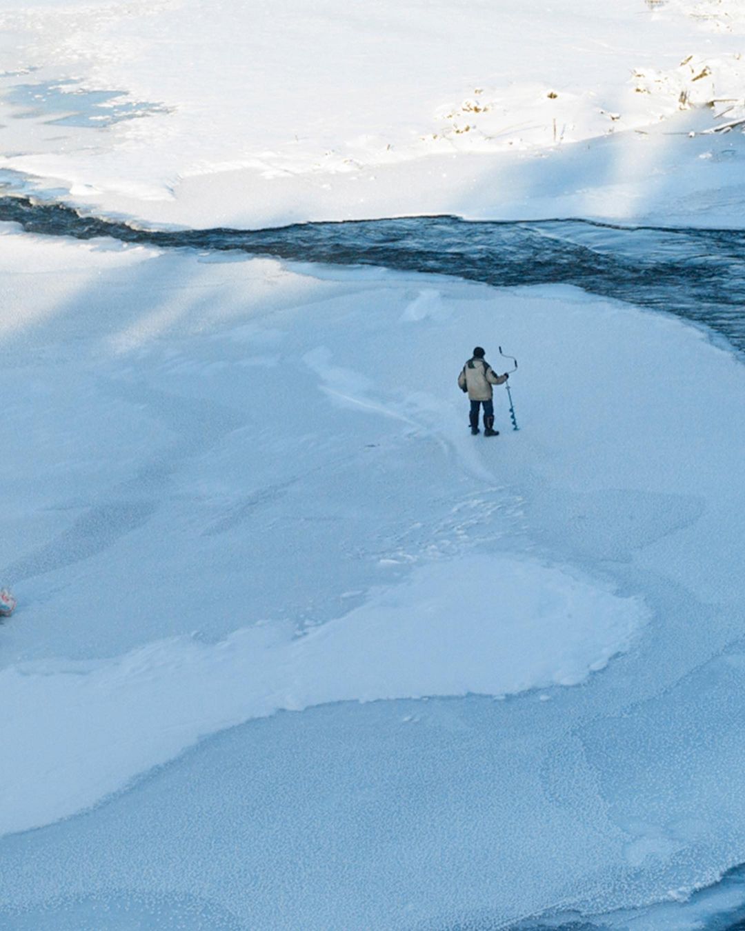 A man ice fishing in winter stands on a frozen river, holding an ice auger. A view from above shows the winter landscape.