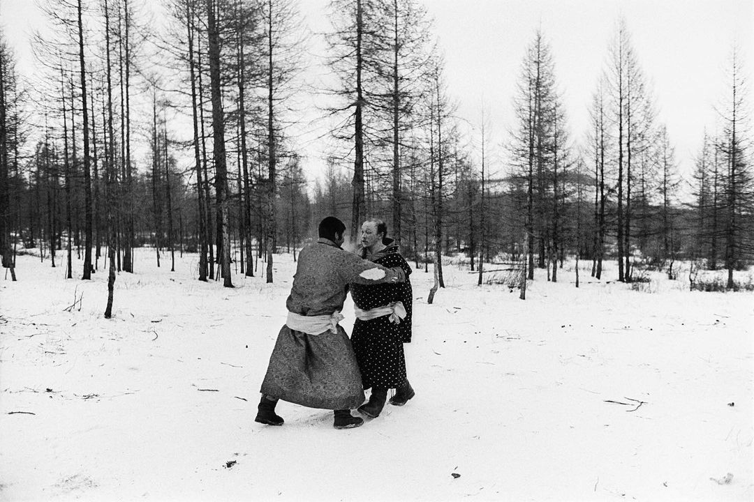 Two adults in traditional garments embrace in a snowy landscape surrounded by tall bare trees in a black and white medium shot.