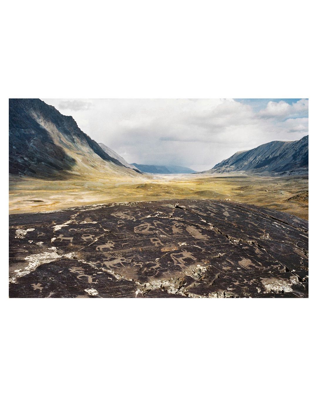 Vast mountain valley with ancient petroglyphs carved into foreground rock, under an overcast sky, creating a scenic vista.