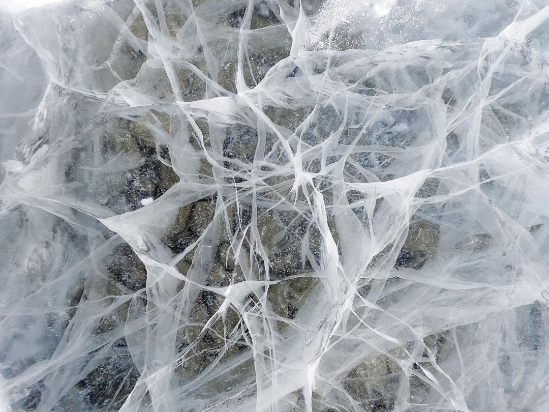 Cracked ice surface with patterns and textures, gray and white tones, creating an abstract and natural visual.