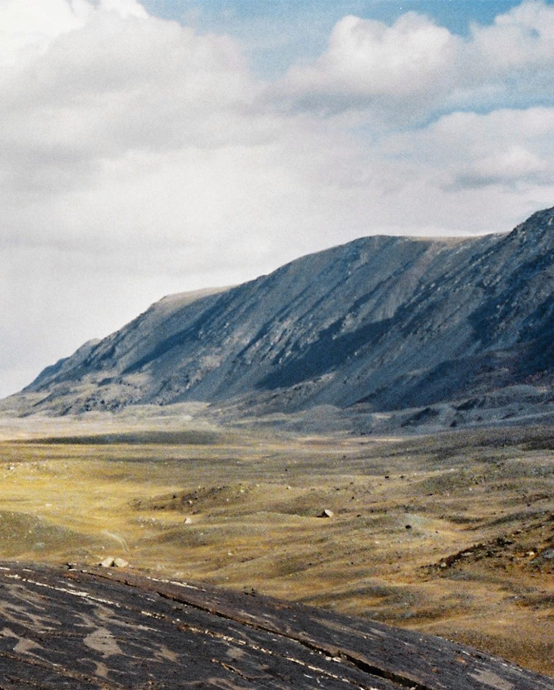 Wide shot of a mountain range and a grassy plain under a cloudy sky. The landscape features a grey mountain, green plains and blue sky.
