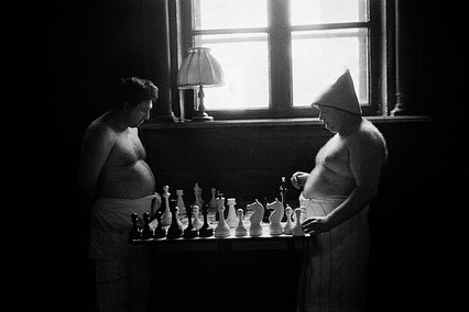 Two men playing chess in a dimly lit room. One man wears a towel and a sauna hat. Natural light enters through a window in the background.
