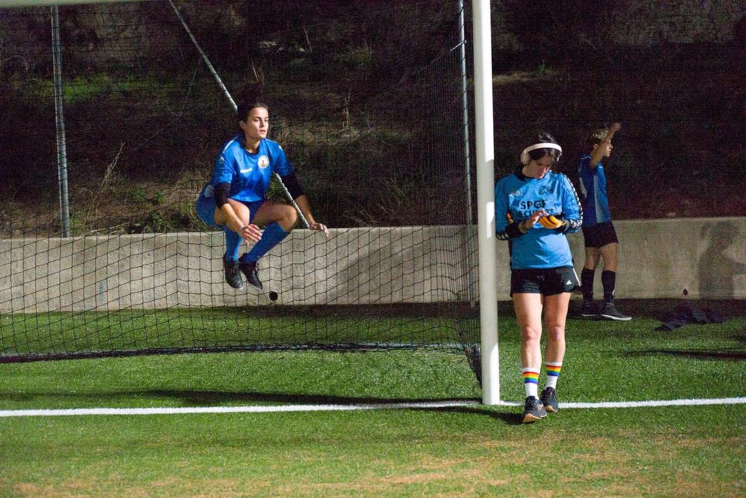 Female athletes on a soccer field at night, one jumping over a net, another standing with rainbow socks, and a third in the background.