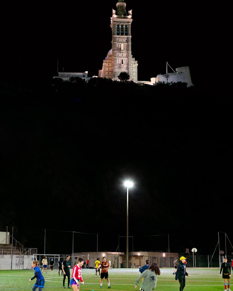 Night view of soccer game in progress on a green field below an illuminated building on a hill; dark sky, bright floodlights.
