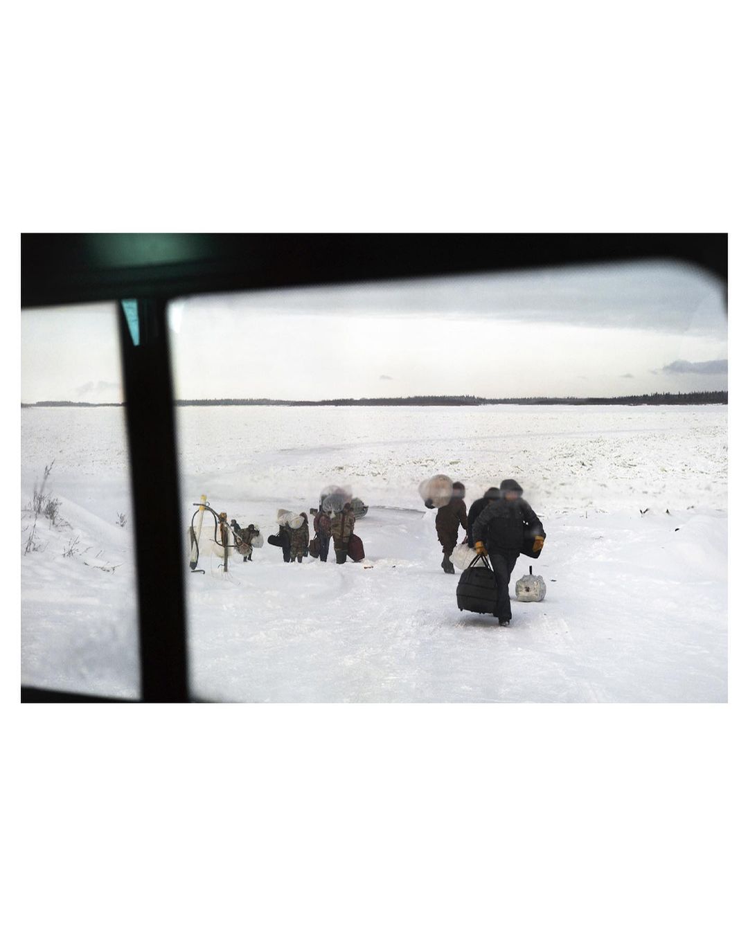 Group of people walk in a snowy landscape, carrying bags, viewed through window; winter scene with overcast sky and bare trees.
