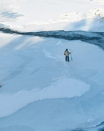 High-angle view of a person standing on a frozen snow-covered river holding an ice auger.