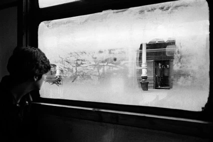 A person looking through a frosted train window at another person standing in a doorway on a platform, captured in black and white.