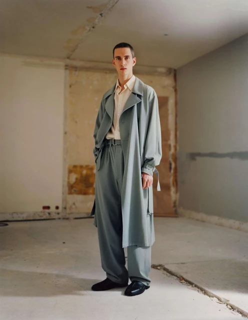An adult male fashion model poses in a blue trench coat and matching pants in a stark indoor environment with distressed walls.