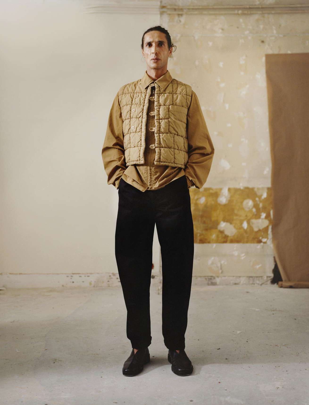 Full-length shot of a single adult male wearing a quilted vest, collared shirt, and dark trousers standing in an indoor studio setting.