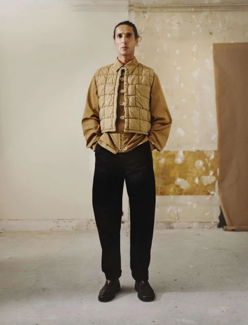 Full-length shot of a single adult male wearing a quilted vest, collared shirt, and dark trousers standing in an indoor studio setting.