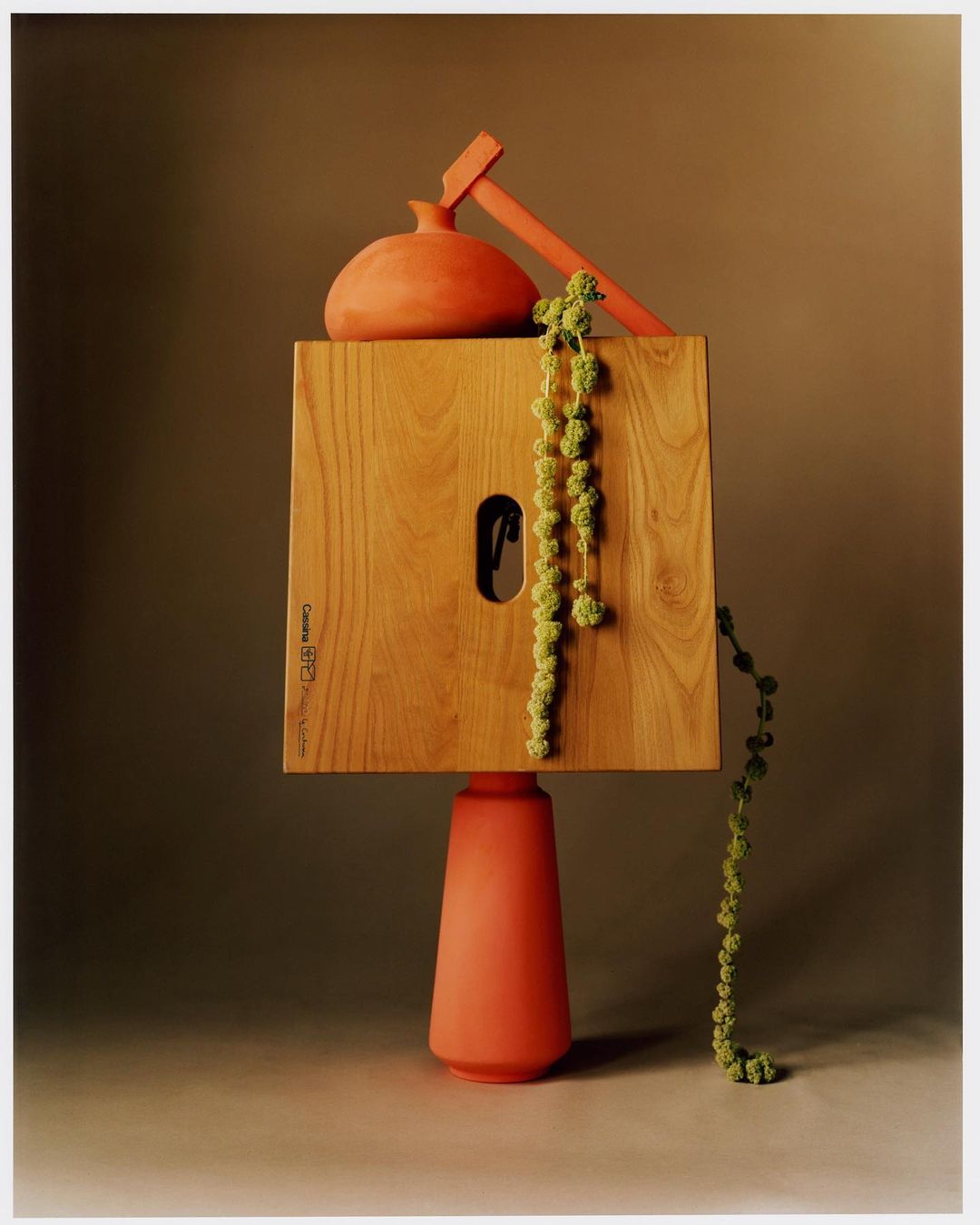 Still life shot with a wooden square, vase, and greenery, showcasing an abstract composition in warm, earthy tones on a neutral backdrop.