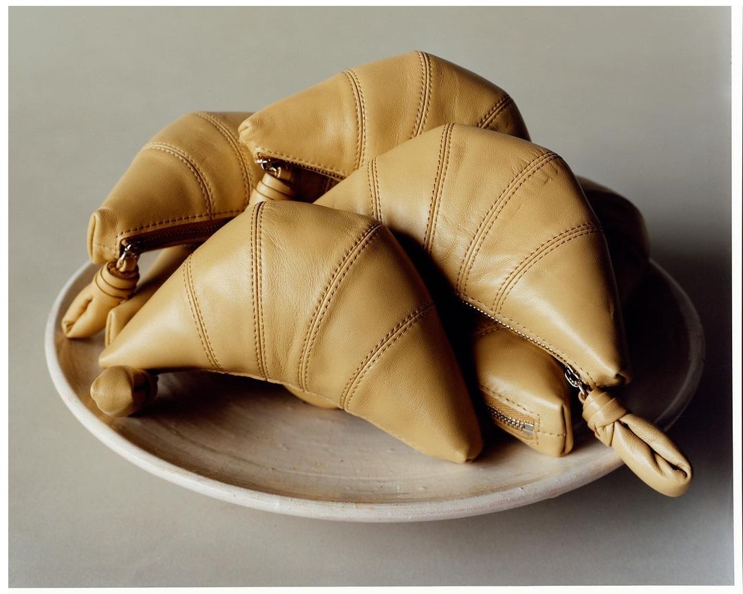 Still life of tan, leather croissant-shaped handbags piled on a beige plate in a studio setting with diffused lighting.