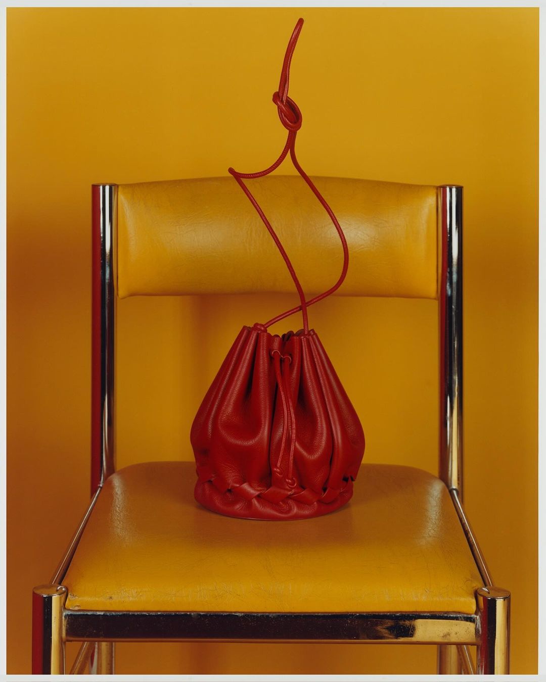 Red leather drawstring pouch bag sits on a yellow leather and chrome chair against a solid yellow background in a studio shot.