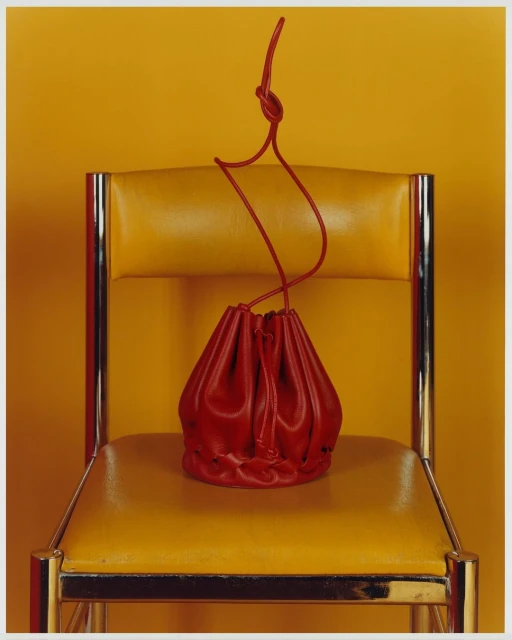 Red leather drawstring pouch bag sits on a yellow leather and chrome chair against a solid yellow background in a studio shot.