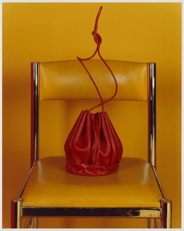Red leather drawstring pouch bag sits on a yellow leather and chrome chair against a solid yellow background in a studio shot.