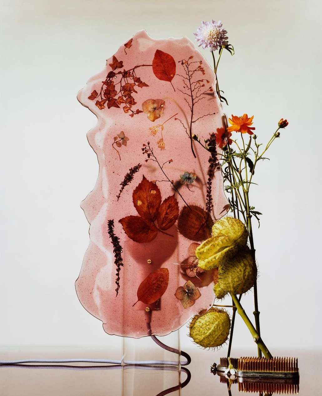 Still life composition featuring an abstract, rose-tinted resin panel with embedded pressed leaves, accented by fresh flowers and vintage pin frog.