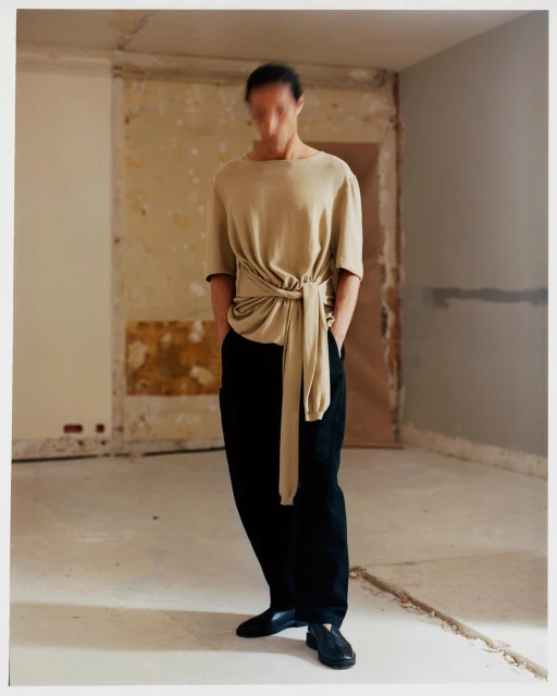 An adult is posing indoors wearing a beige top with tie and black trousers, with black shoes, against a wall with neutral colors.