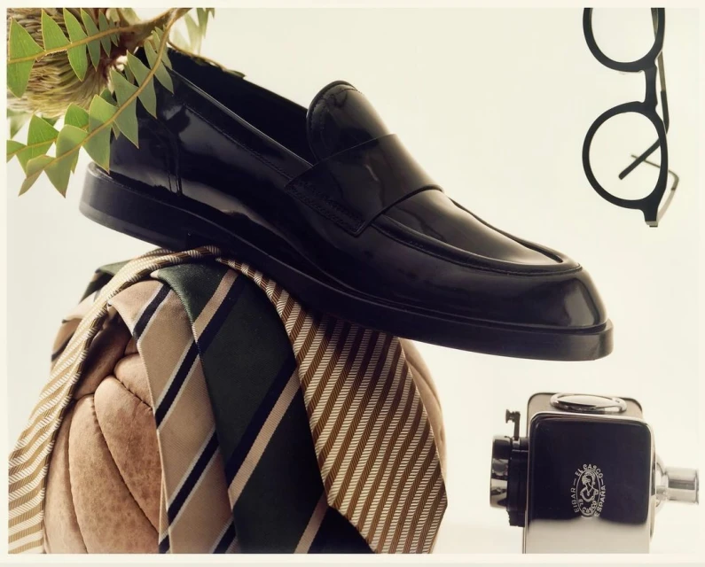 A black leather penny loafer rests atop a collection of striped neckties, complemented by eyeglasses and a vintage-style sharpener.