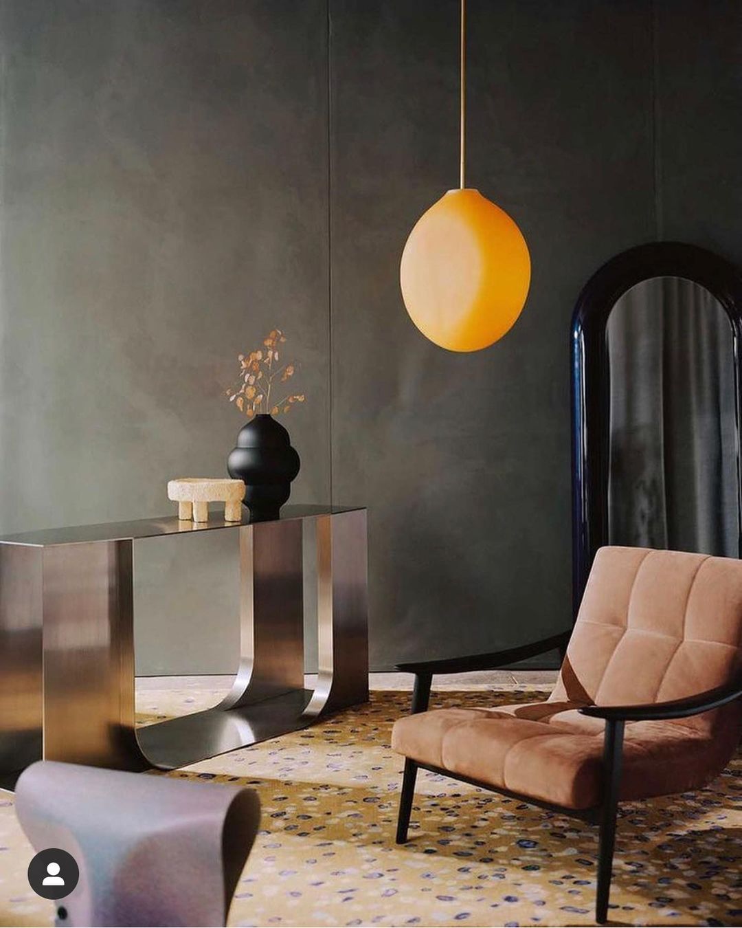 Living room interior featuring a brown armchair, brushed metal console table, modern black vase, and an orange pendant light.