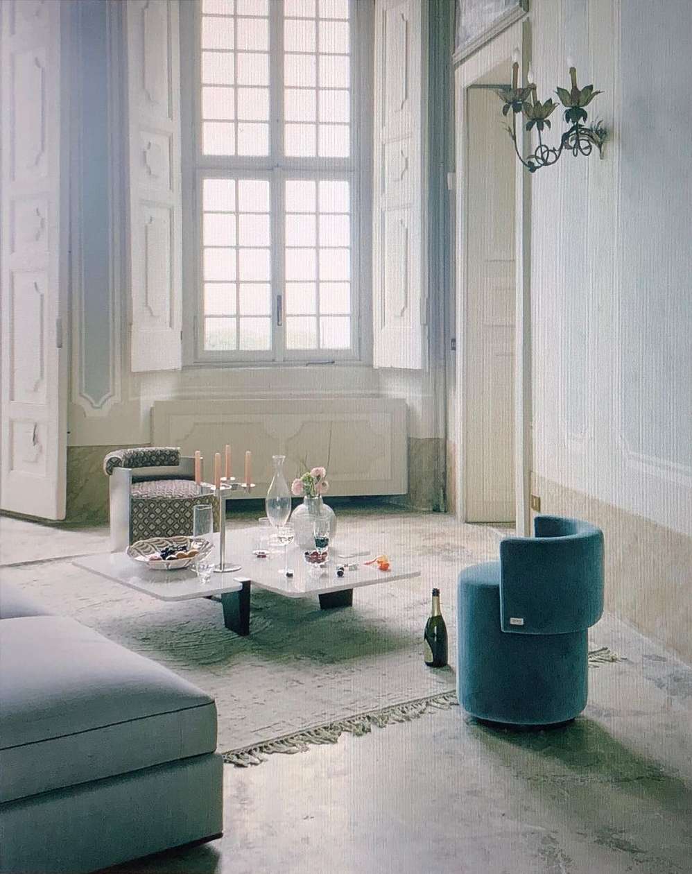 Brightly lit living room with a teal sofa, rounded ottoman, marble coffee table, and decorative objects, creating a calm and elegant interior.