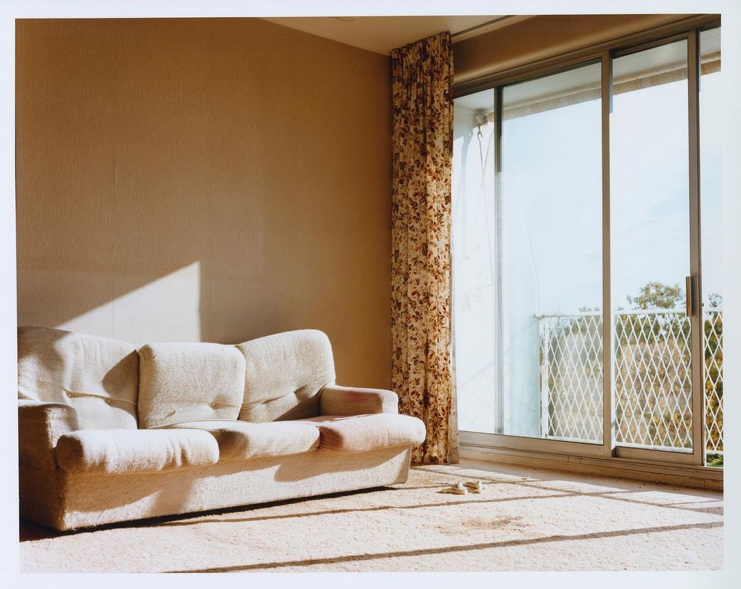 A brightly lit, empty living room interior with an off-white couch, floral curtains, and a sliding glass door leading to a balcony.