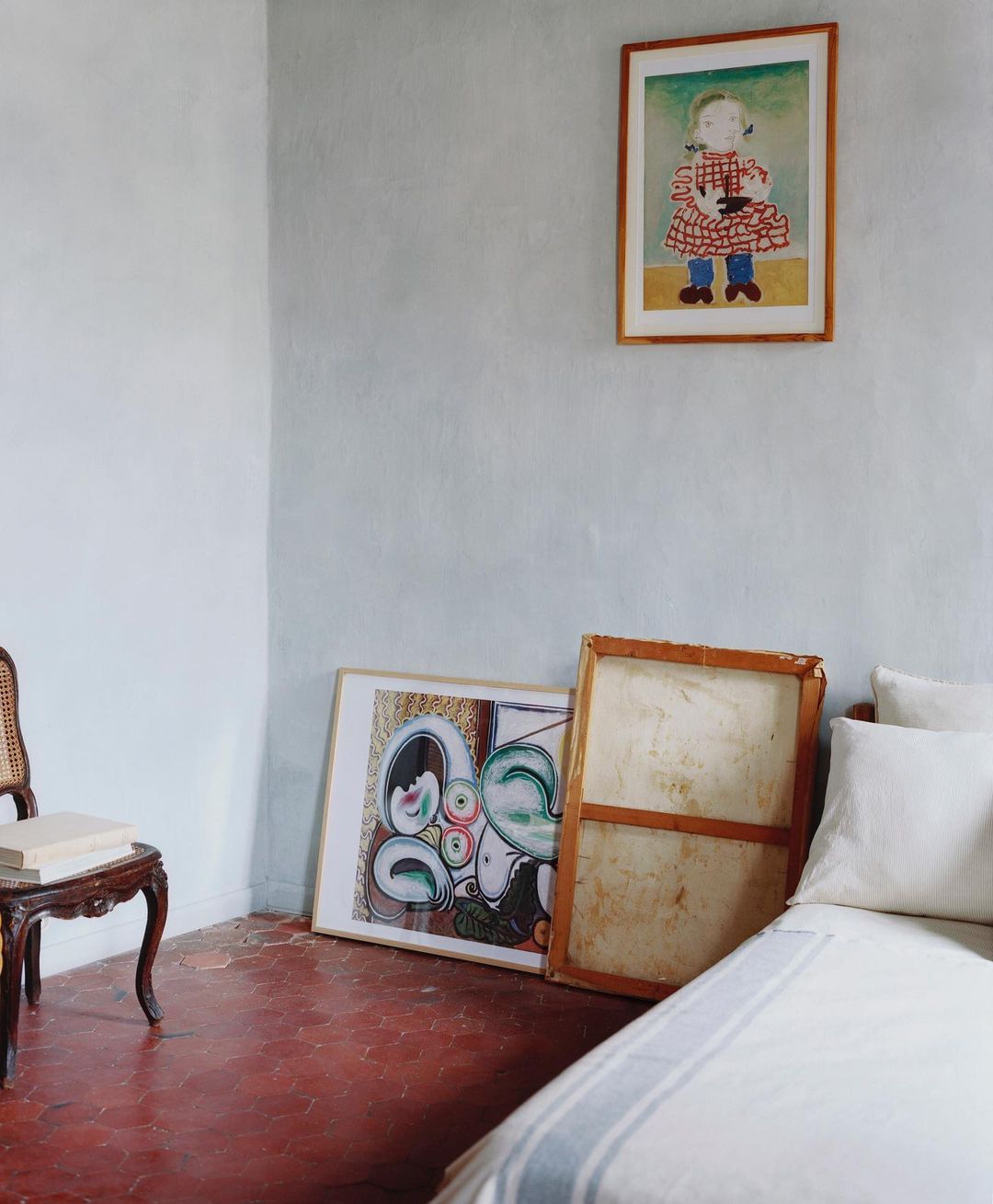 Interior scene features framed drawings on a light blue wall, a vintage cane chair, and a partially made bed with pillows.