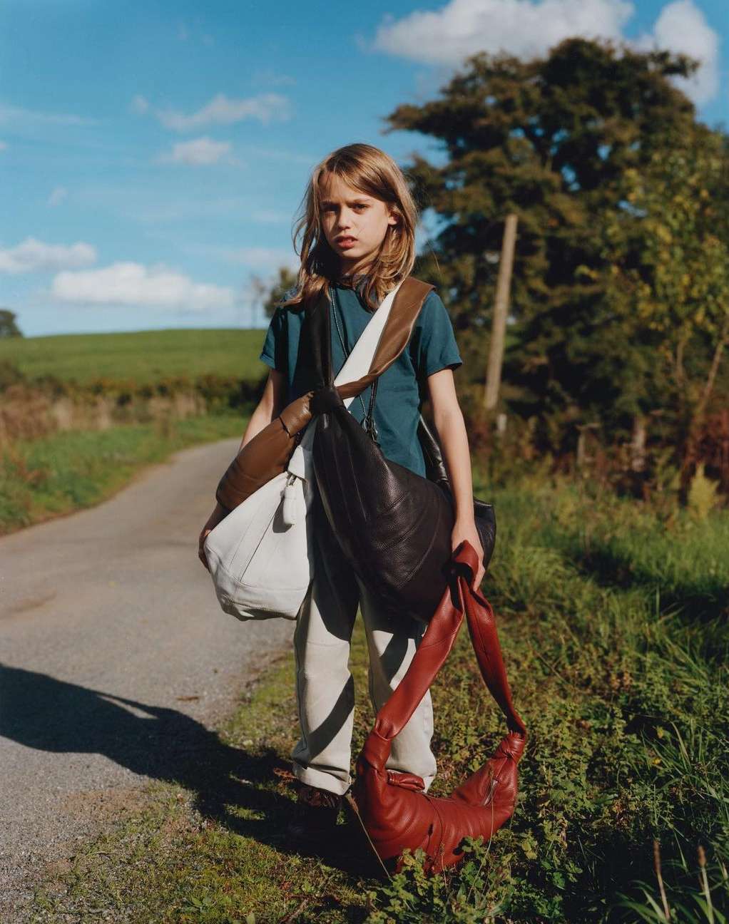 Young person stands on rural road carrying multiple shoulder bags, with grass, trees, and blue sky in background on a sunny day.