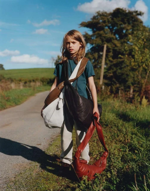 Young person stands on rural road carrying multiple shoulder bags, with grass, trees, and blue sky in background on a sunny day.