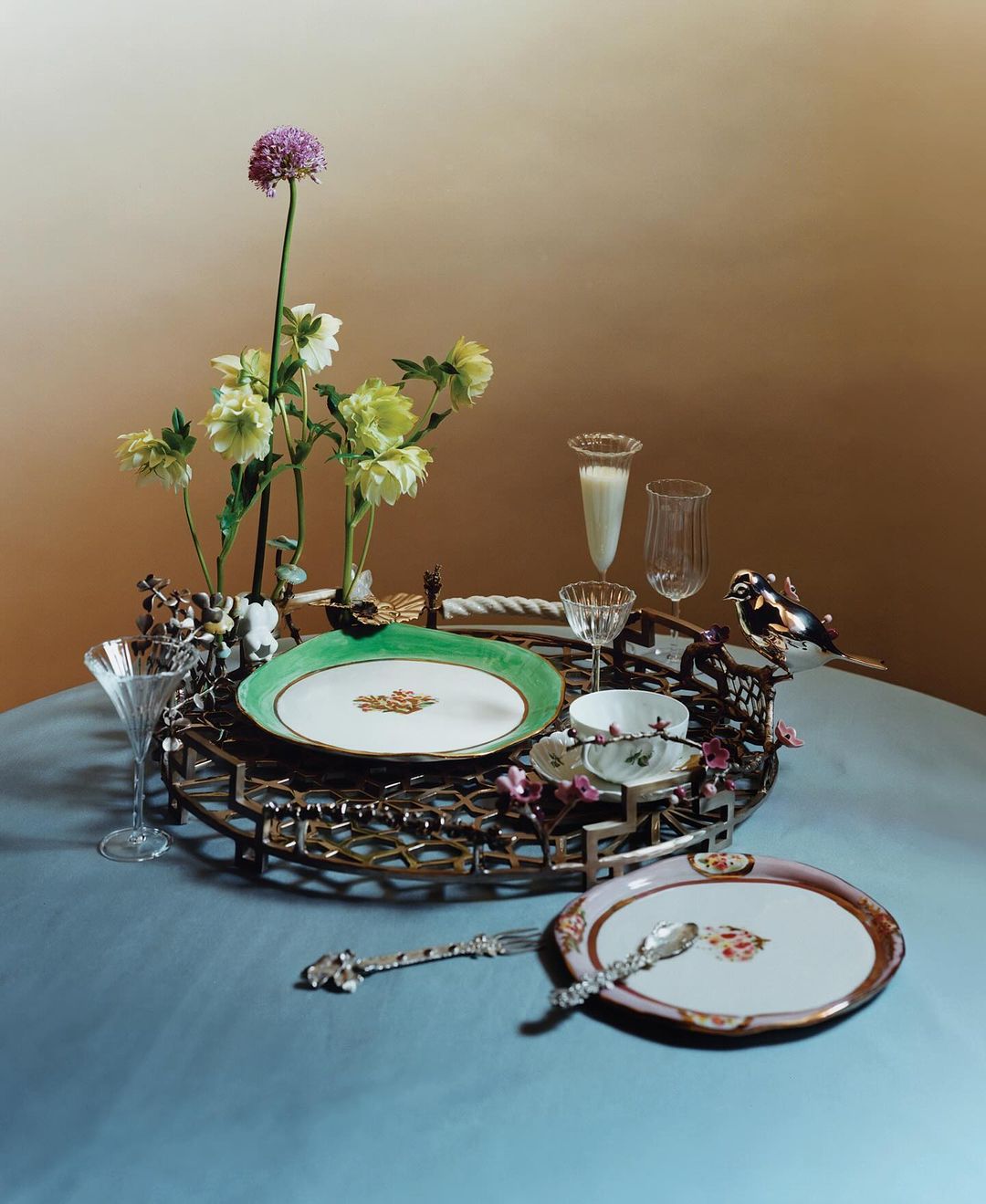 Still life shot of a table setting featuring fine china, crystal glasses, flowers, and silverware against a brown to blue background.