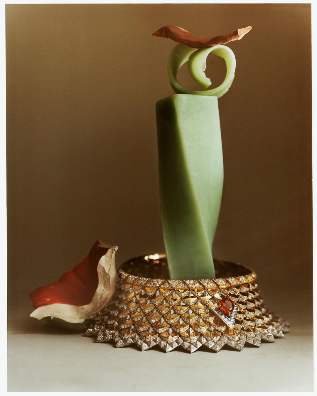 Still life shot of a chunky gold necklace, adorned with a red gemstone, with a green sculpture and a brown leaf on top.