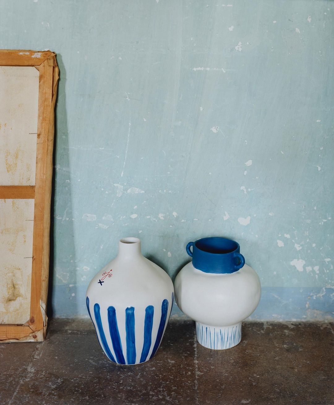 Two decorative vases are placed on a stone surface against a blue textured wall, one with blue stripes and a painted canvas on the side.