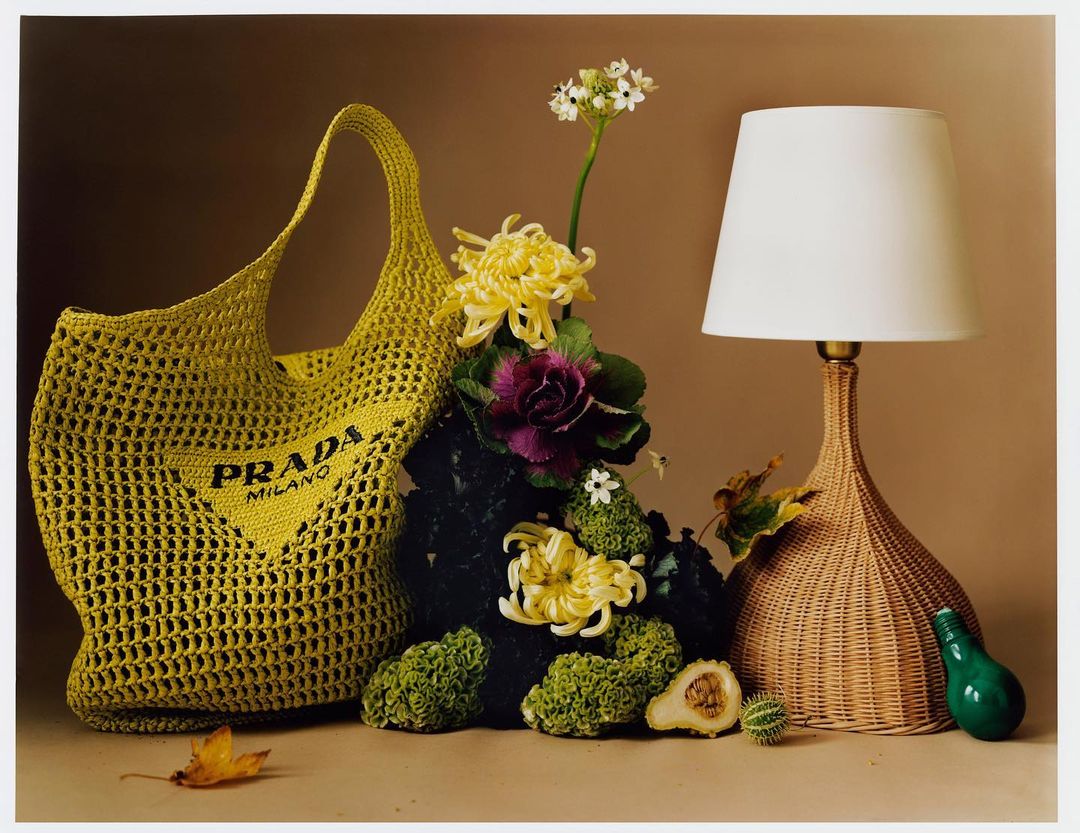 A yellow crochet Prada Milano tote bag next to a lamp with flowers and foliage in a still life composition on a beige background.
