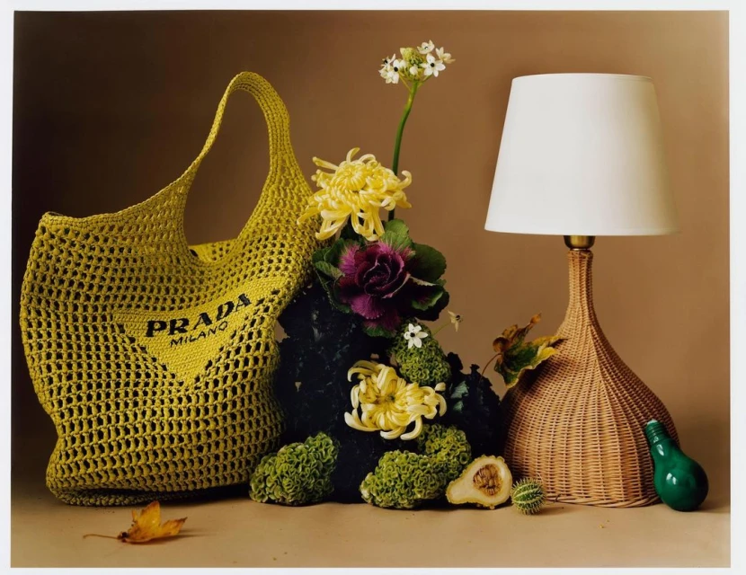 A yellow crochet Prada Milano tote bag next to a lamp with flowers and foliage in a still life composition on a beige background.