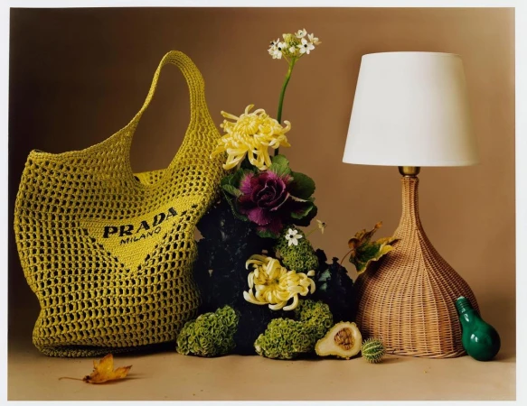 A yellow crochet Prada Milano tote bag next to a lamp with flowers and foliage in a still life composition on a beige background.