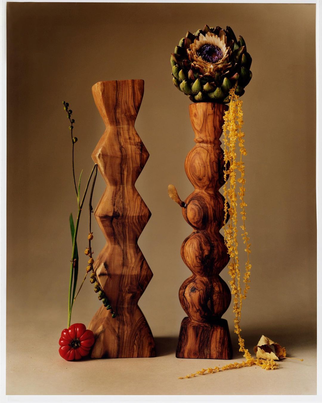 Still life with wooden sculptures, artichoke flower, and pepper in a warm-toned studio environment featuring artful lighting and composition.