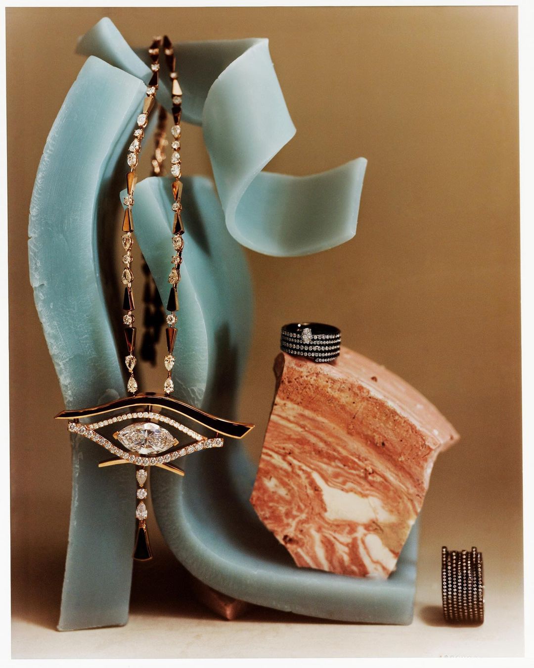 Still life shot of a gold eye-shaped pendant necklace with diamonds, a ring, and stone fragments against a neutral and turquoise background.