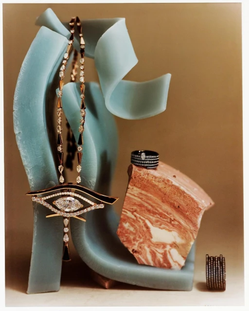 Still life shot of a gold eye-shaped pendant necklace with diamonds, a ring, and stone fragments against a neutral and turquoise background.