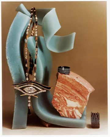 Still life shot of a gold eye-shaped pendant necklace with diamonds, a ring, and stone fragments against a neutral and turquoise background.