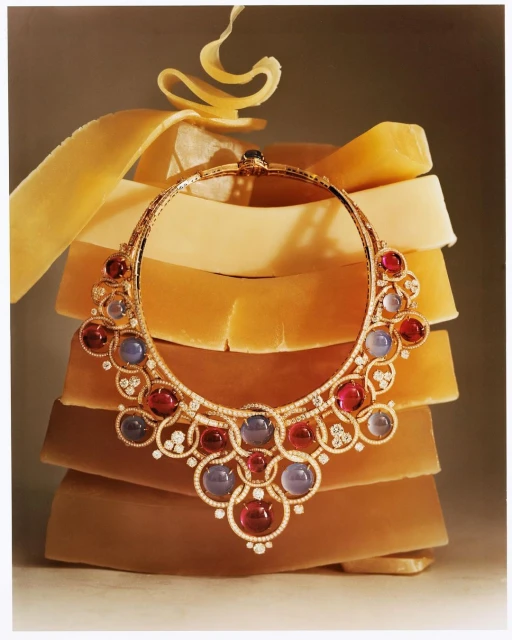 Elegant gold necklace featuring gemstones and diamonds displayed on stacked blocks with a yellow curl element on top in a studio setting.