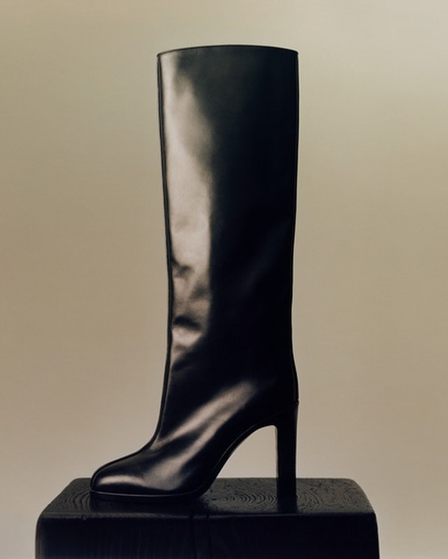 Black knee-high heeled boot displayed on a black wooden block against a neutral backdrop in a studio setting. Fashion photography.